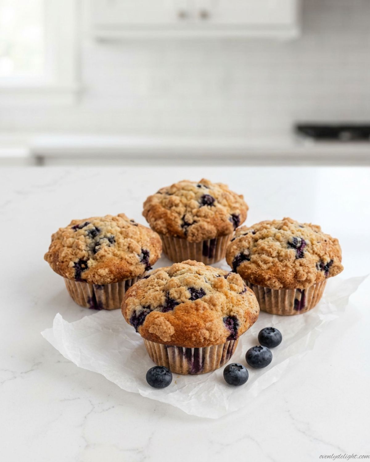 Top-down view of four bakery-style blueberry muffins arranged on a clean white countertop, featuring golden domed tops with juicy blueberries, set against a bright luxury kitchen background with shallow depth of field.